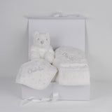 A baby gift hamper containing a white hooded towel, a white teddy bear, a white bandana bib, a white sleeping bag, and a white bath mitt, all displayed in a white box with a keepsake box and a gift card.